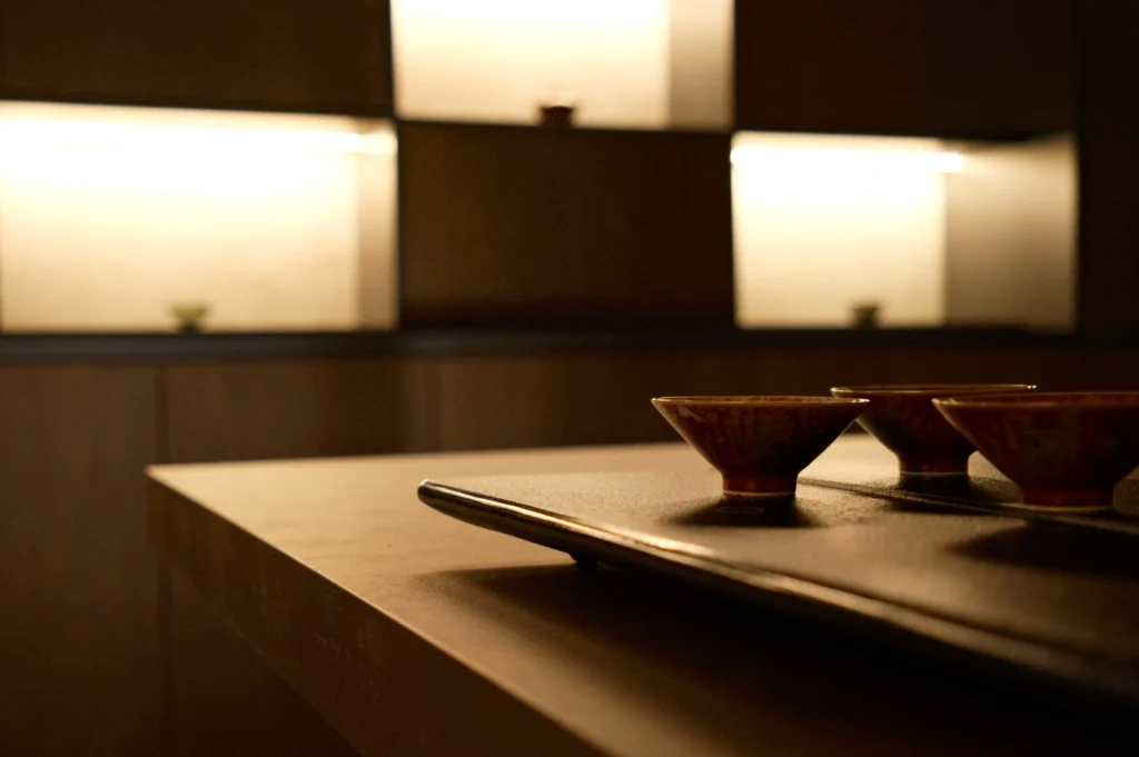 A minimalist wooden table with small, decorative bowls on a tray. Soft ambient lighting illuminates shelves in the background, creating a serene atmosphere.