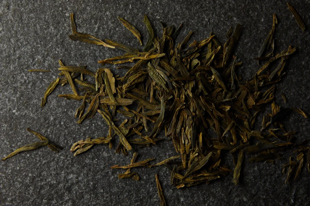 A pile of dried green tea leaves scattered on a textured dark gray stone surface, showcasing their elongated shape and earthy green hue.
