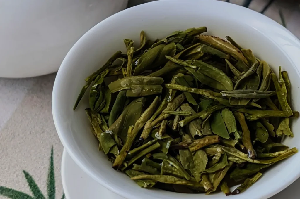 A close-up of a white bowl filled with wet green tea leaves, suggesting freshness. The background includes a hint of a white teapot and tablecloth.