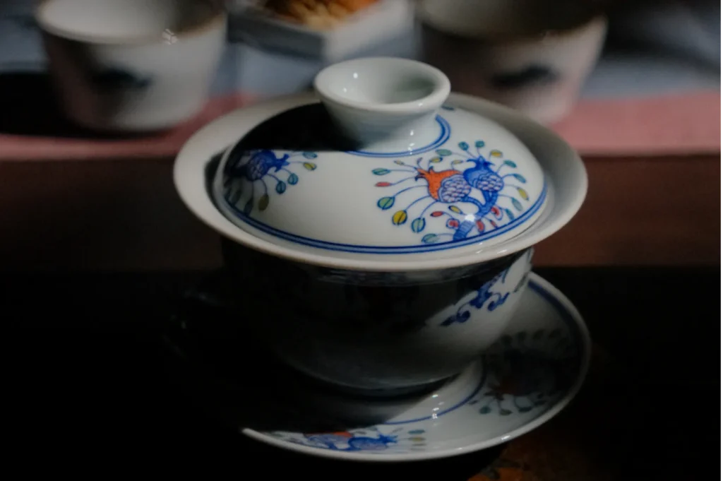 A traditional Chinese tea cup with a lid sits on a matching saucer, adorned with blue and orange floral designs, creating a serene, elegant atmosphere.