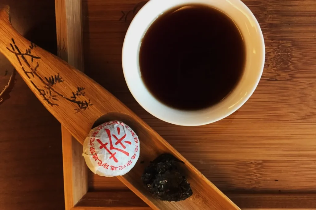 White teacup with dark tea on a wooden tray, beside a bamboo scoop holding a wrapped tea ball and a small piece of loose leaf tea, evoking a calm, traditional vibe.