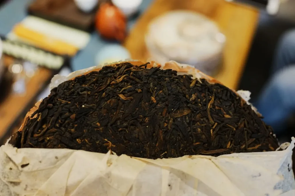 Close-up of a round, compressed tea cake wrapped in paper, showing dark brown tea leaves. The background is blurred, suggesting a casual tea setting.