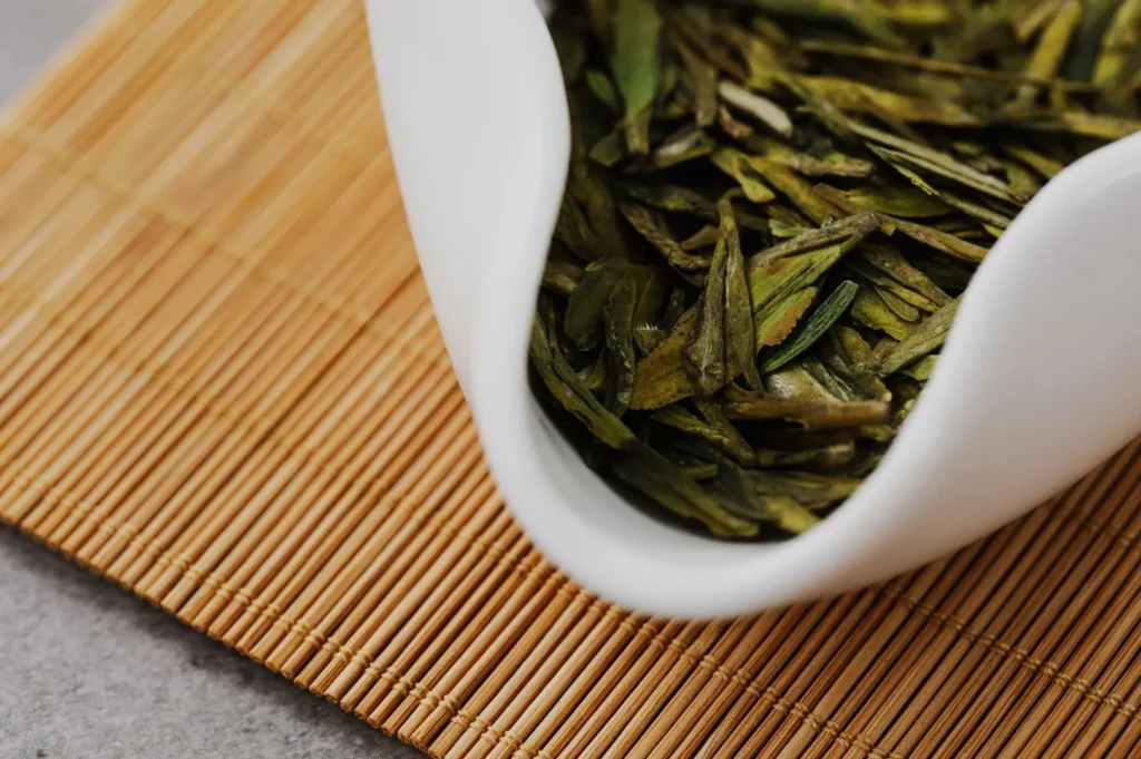 A white bowl filled with dried green tea leaves sits on a bamboo mat, conveying a sense of freshness and tranquility in a minimalist setting.