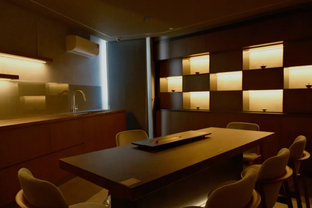 Dimly lit modern dining room with a wooden table, chairs, and minimalist shelving. Soft ambient lighting creates a cozy, intimate atmosphere.