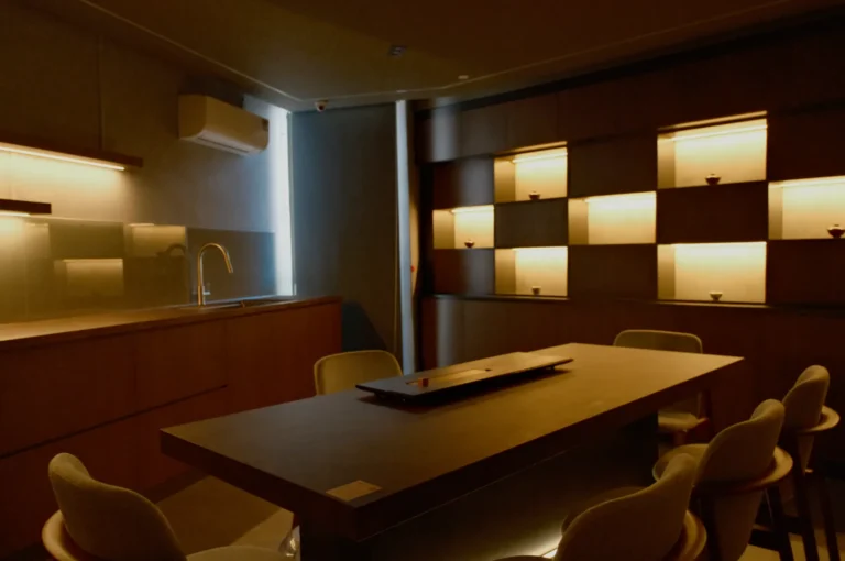 Dimly lit modern dining room with a wooden table, chairs, and minimalist shelving. Soft ambient lighting creates a cozy, intimate atmosphere.
