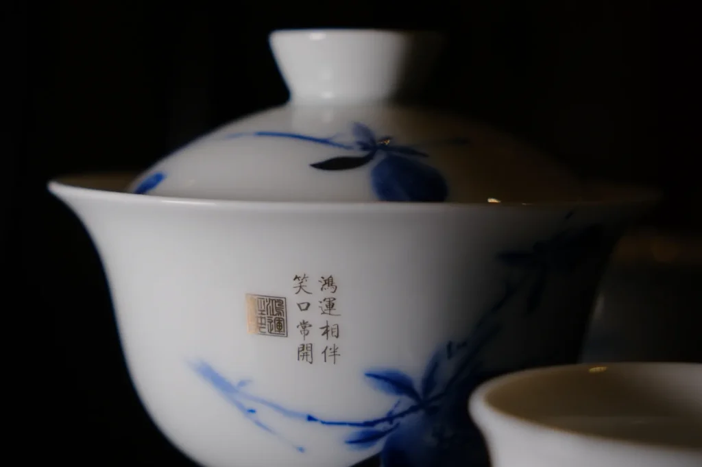 A white porcelain teacup with a fitted lid displays delicate blue dragonfly motifs and Chinese characters. The scene feels serene and traditional.