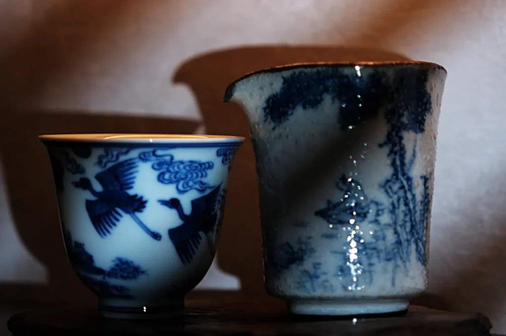 Two blue and white porcelain cups with intricate designs. The left cup features birds in flight, while the right has a serene landscape scene.
