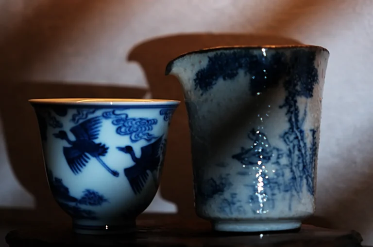 Two blue and white porcelain cups with intricate designs. The left cup features birds in flight, while the right has a serene landscape scene.