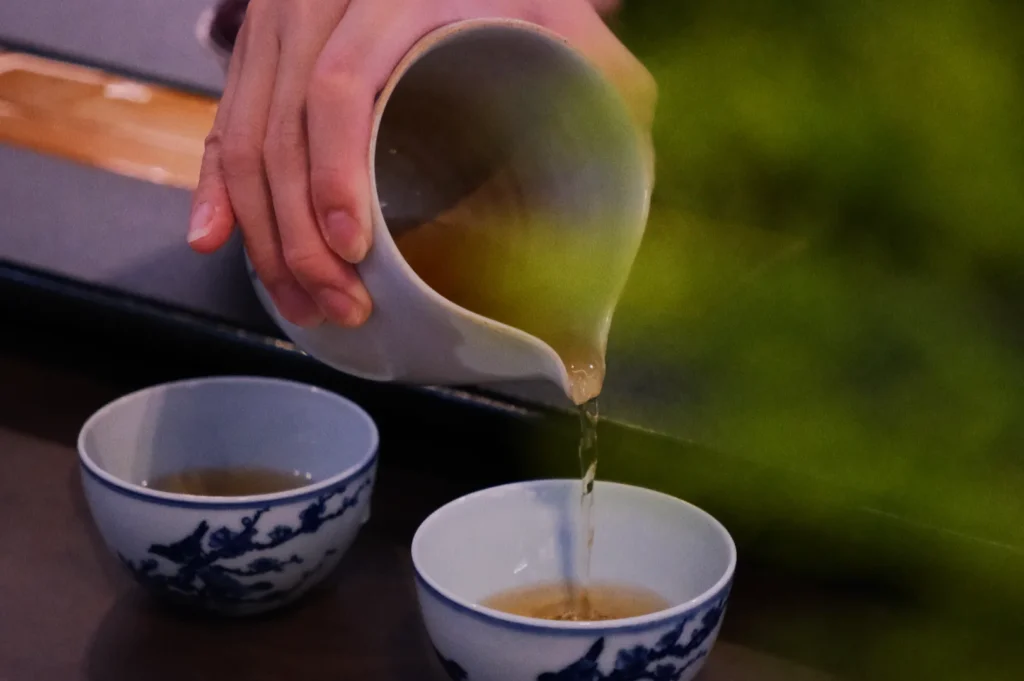 A hand pours tea from a white ceramic pot into a small cup with blue floral designs. Another filled cup is nearby. The scene is calm and inviting.