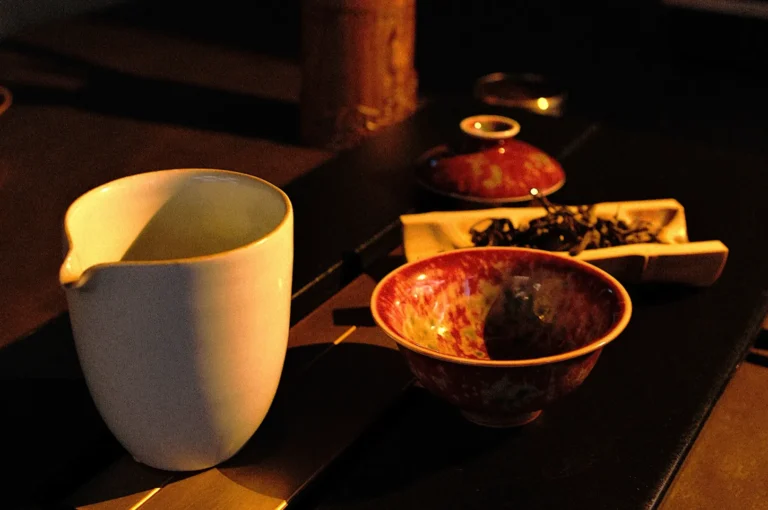 Warmly lit still life of a white pitcher, a colorful ceramic bowl, and a small dish with tea leaves, all set on a dark wooden surface.