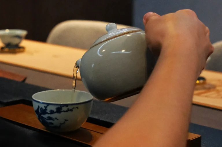 A hand pours tea from a ceramic teapot into a decorated cup on a wooden tray. The scene conveys a sense of calm and tradition.