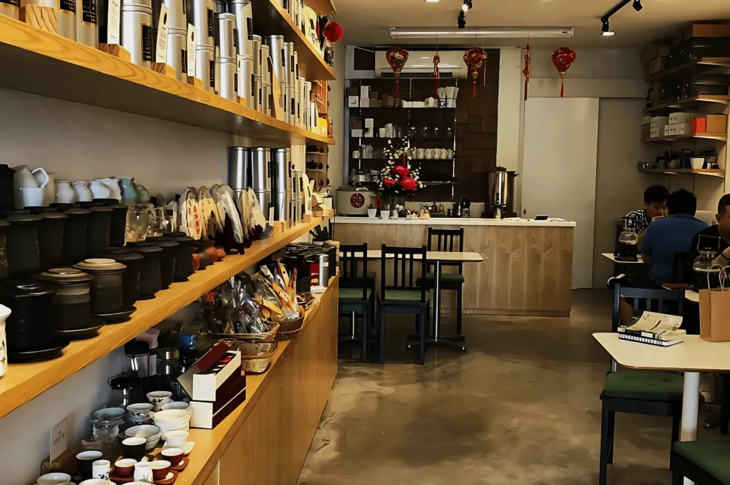 Cozy tea shop interior with wooden shelves lined with tea sets and accessories. Tables at the back accommodate patrons. Red lanterns add festive decor.