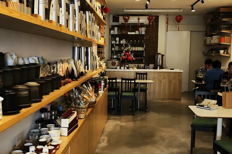Cozy tea shop interior with wooden shelves lined with tea sets and accessories. Tables at the back accommodate patrons. Red lanterns add festive decor.