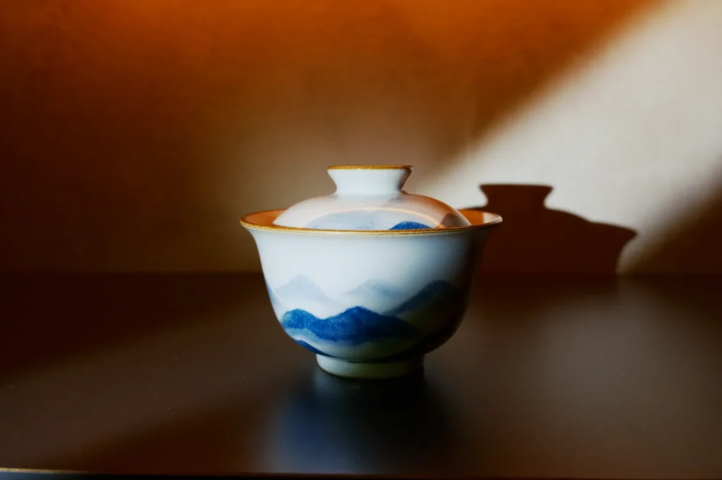 A delicate ceramic tea cup with a lid painted with blue mountains sits on a dark surface, lit softly from the side, casting a gentle shadow.
