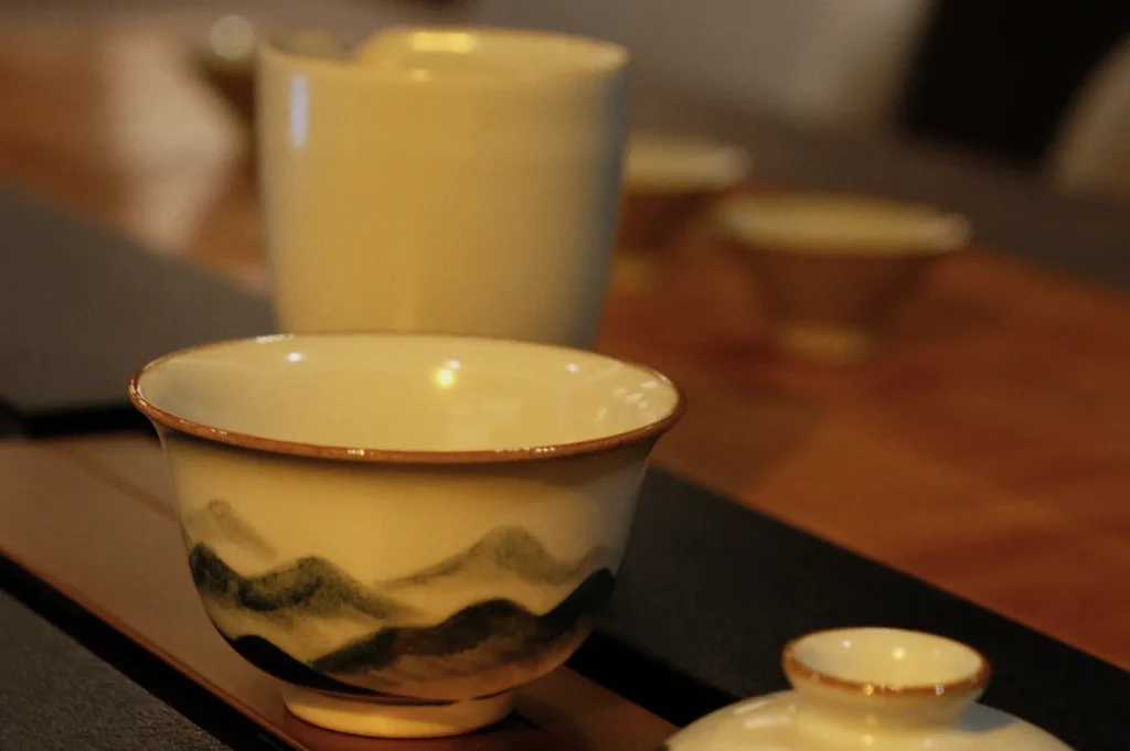 Close-up of an elegant, white tea cup with a painted mountain design and gold rim, surrounded by other ceramic cups on a wooden table, conveying a serene atmosphere.
