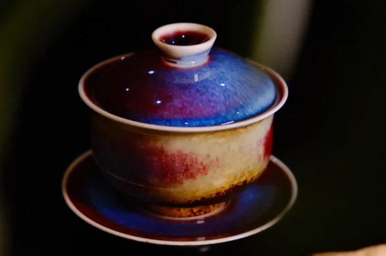 A colorful ceramic teacup with a lid sits on a matching saucer. The glaze blends deep blue, red, and cream hues, creating a vibrant, artistic look.
