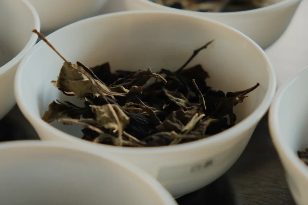 A close-up of a white bowl filled with dried tea leaves. The dark, crinkled leaves suggest an earthy aroma, set on a wooden table.