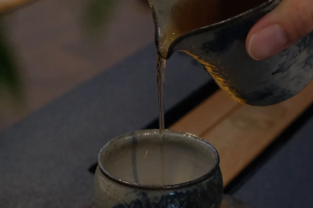 A hand pours hot tea from a rustic, ceramic pitcher into a matching cup. The setting is cozy and warm, suggesting a peaceful, calming moment.