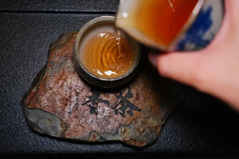 A hand pours golden tea from a ceramic pot into a cup resting on a textured stone with black Asian script, creating a serene and mindful atmosphere.