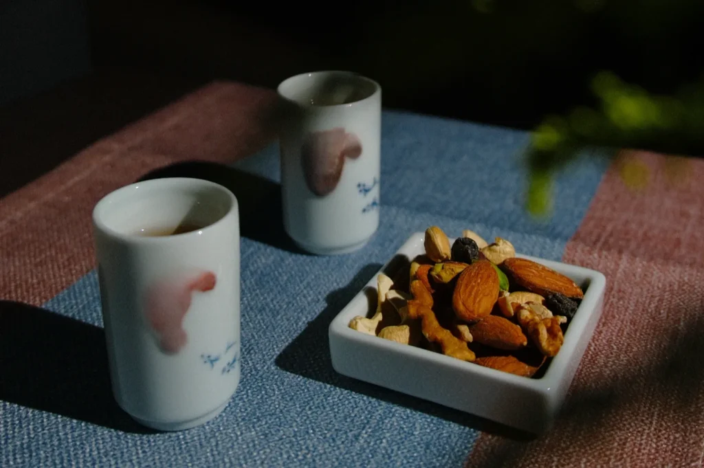 Two ceramic cups on a blue and pink tablecloth, partially filled, beside a square dish of mixed nuts. The setting is calm and intimate.