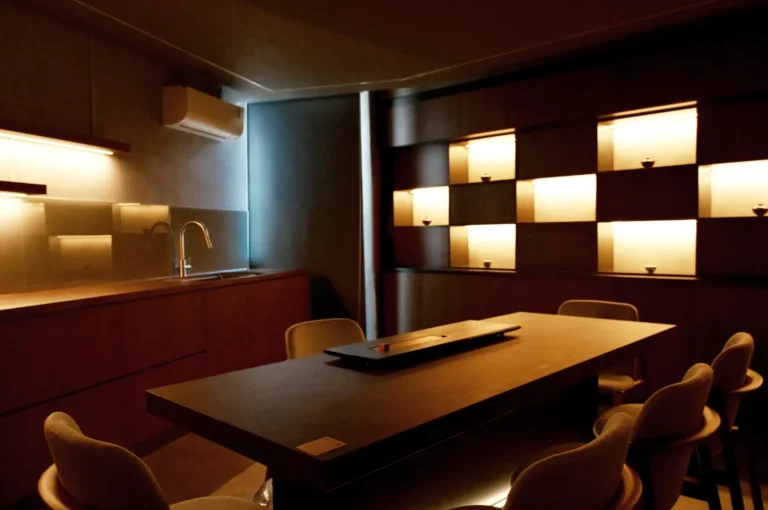 Dimly lit modern kitchen with a sleek counter and chairs. Subtle under-cabinet lighting highlights a sink. Backlit shelving softly illuminates pottery. Cozy and sophisticated ambiance.