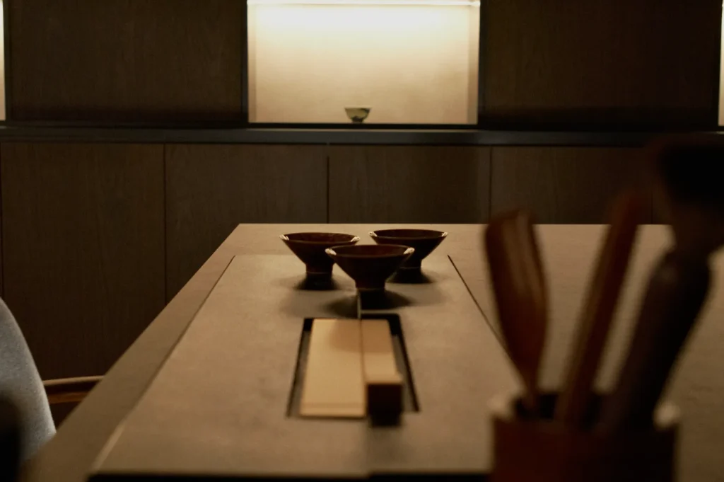 Minimalist setting with a wooden table featuring four small bowls. Softly lit shelves in the background lend a serene, contemplative atmosphere.