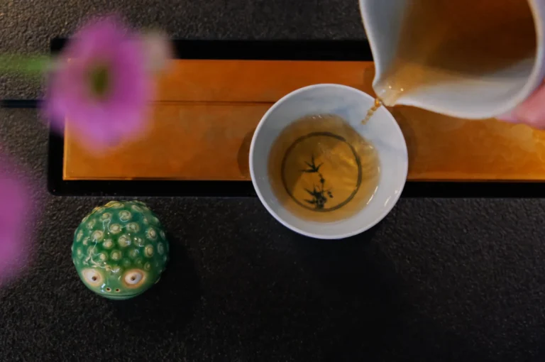 Tea is being poured into a white cup with a leaf design, placed on a yellow mat. A green ceramic figurine and a blurred pink flower are nearby. Calm atmosphere.