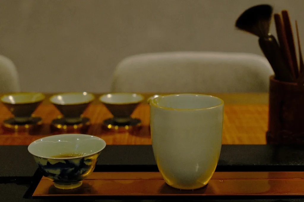 Ceramic cups are arranged on a wooden table with a bamboo tea whisk in the background. The scene conveys a calm, traditional tea setting.