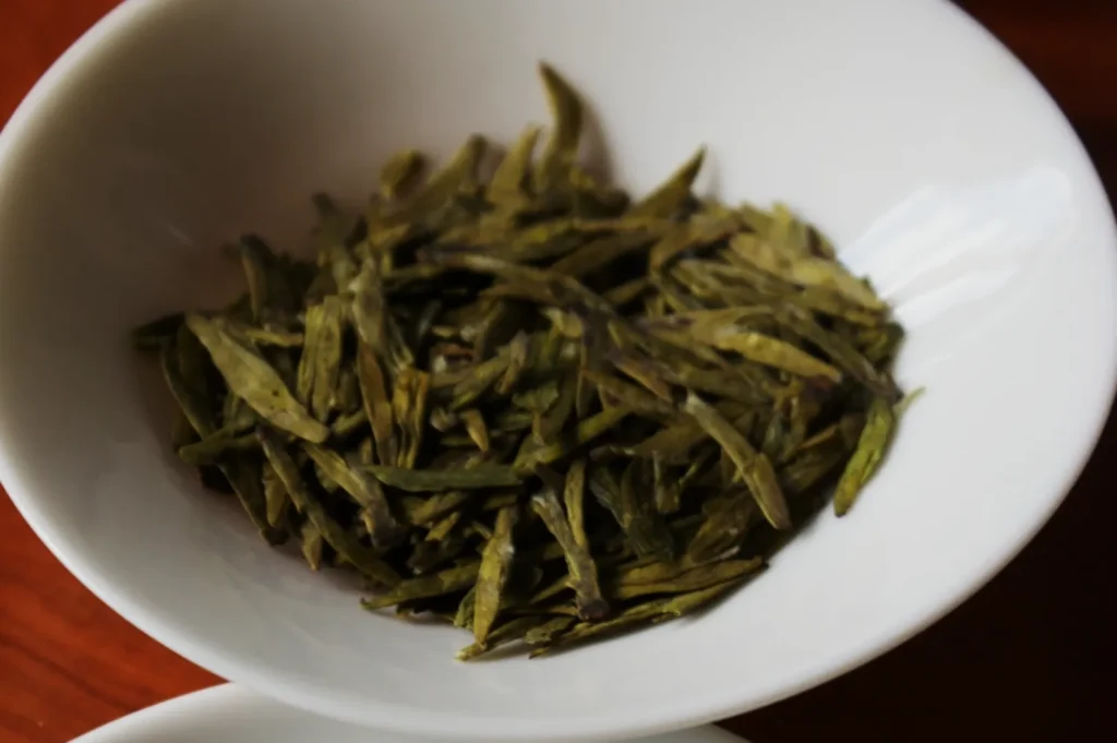 A close-up of dried green tea leaves in a white bowl on a wooden surface. The leaves appear flat and elongated, conveying a sense of freshness.
