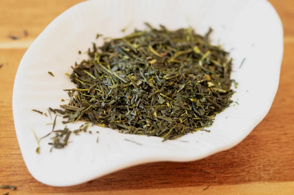 A white dish holds dried green tea leaves, showcasing their rich, earthy texture. The wooden surface underneath adds a warm, natural tone.