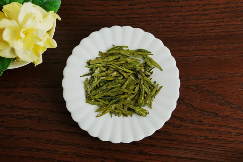 A white scalloped plate with dried green tea leaves sits on a wooden table. A yellow flower in a vase is partially visible on the left. The image conveys freshness.