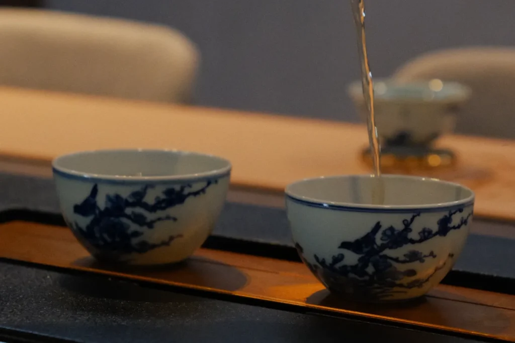 Two porcelain cups with blue floral designs are placed on a wooden tray. Tea is being poured into one cup. The setting is calm and cozy.