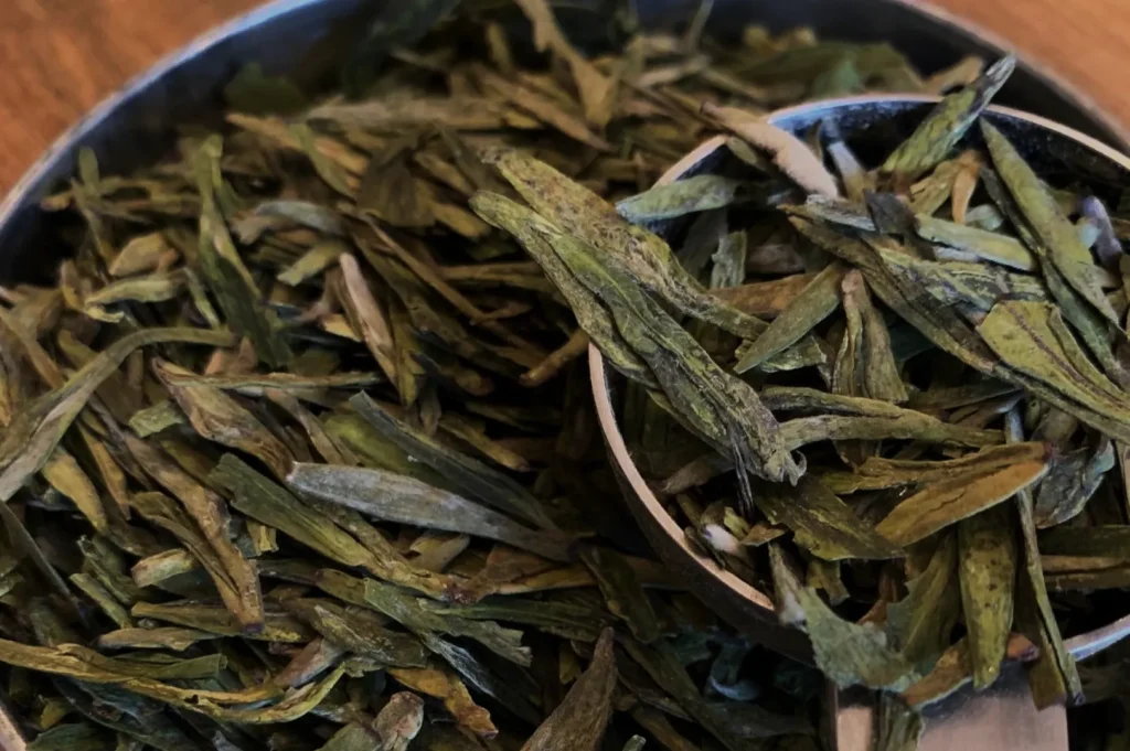 Close-up of a silver scoop filled with long, flat green tea leaves, resting on a pile of similar leaves. The earthy tones convey freshness.