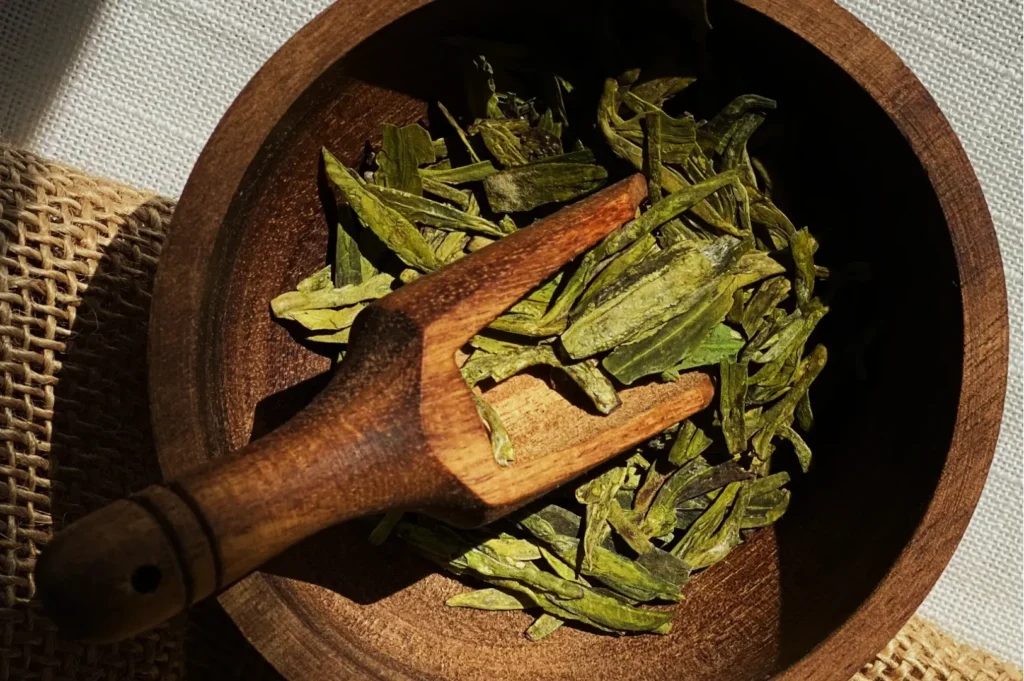 A wooden bowl filled with dried green tea leaves, accompanied by a small wooden scoop. Sunlight casts gentle shadows, evoking a natural, earthy tone.