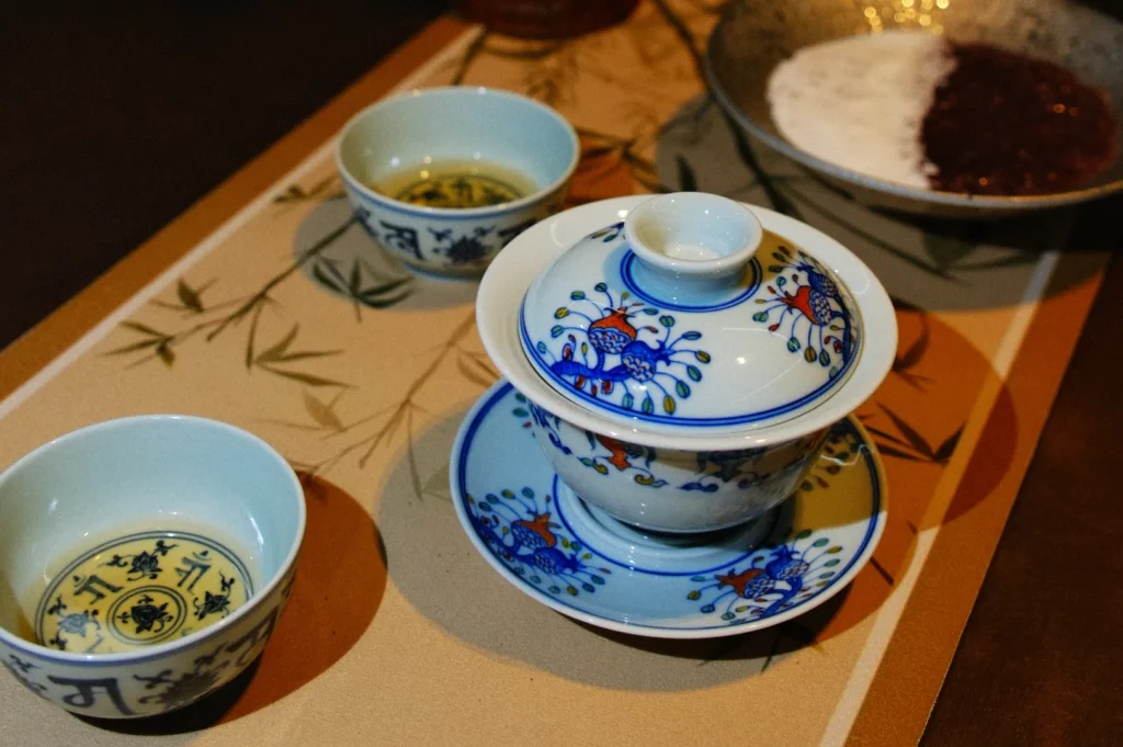 Porcelain tea set with intricate blue and red floral designs on a bamboo-patterned mat. Nearby are two tea cups and a bowl with white and red paste.