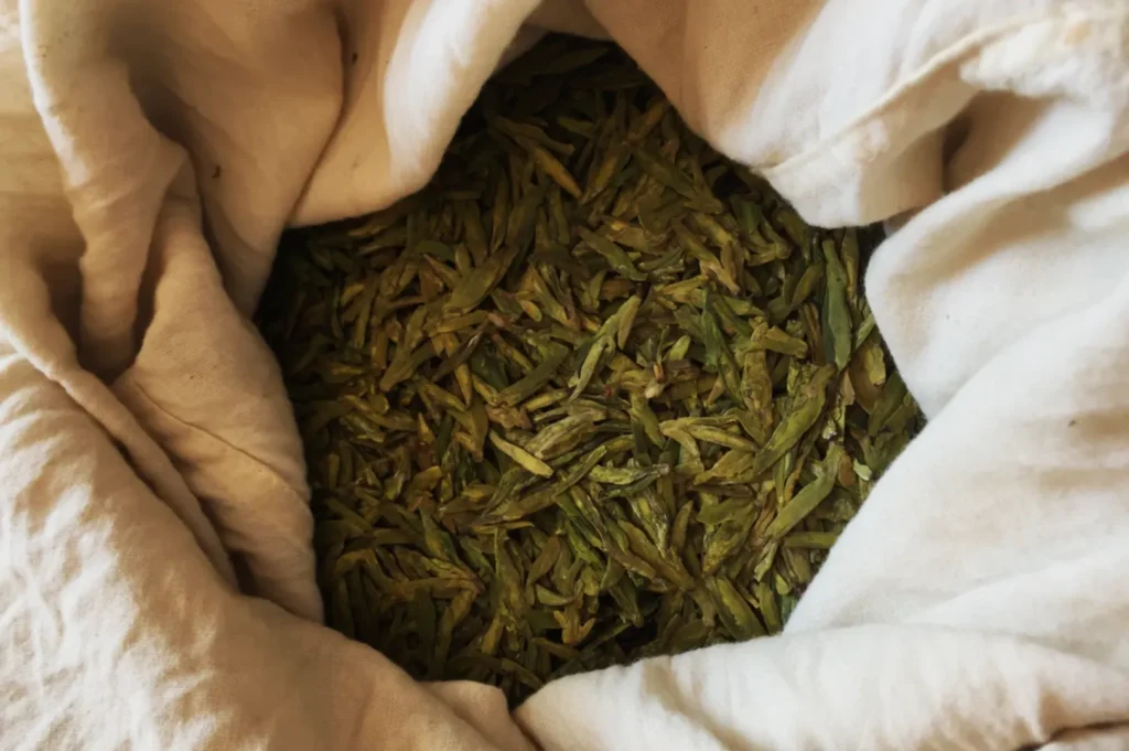 A cloth bag filled with dried green tea leaves, showcasing their vibrant color and texture. The setting suggests freshness and natural simplicity.