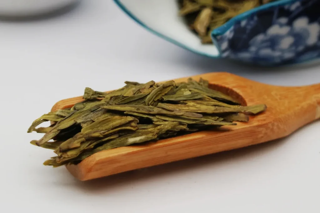 A wooden scoop holds dry green tea leaves, with a decorative blue and white ceramic bowl partially visible in the background. The tone is calm and earthy.