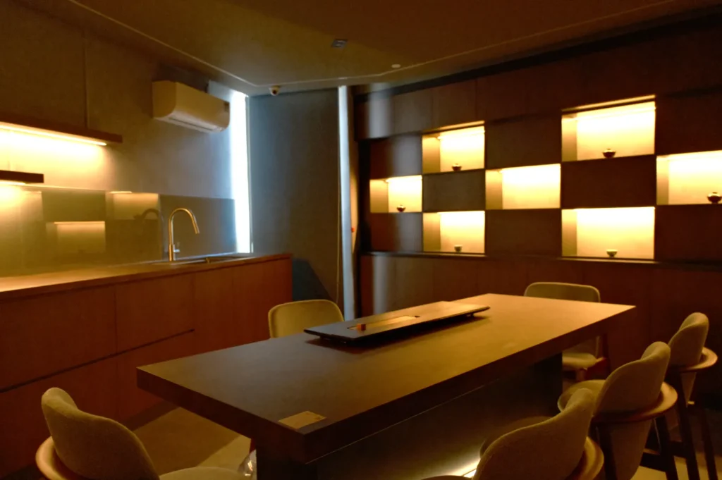 Dimly lit modern dining area featuring a long wooden table with chairs, minimalist decor, and warm backlit shelves displaying small objects. Cozy ambiance.