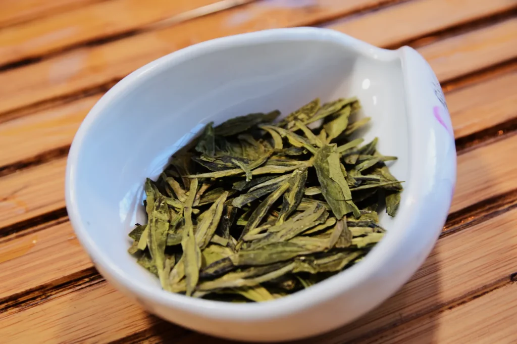 A white ceramic bowl filled with dried green tea leaves sits on a wooden surface. The leaves are flat and elongated, conveying a fresh and natural tone.