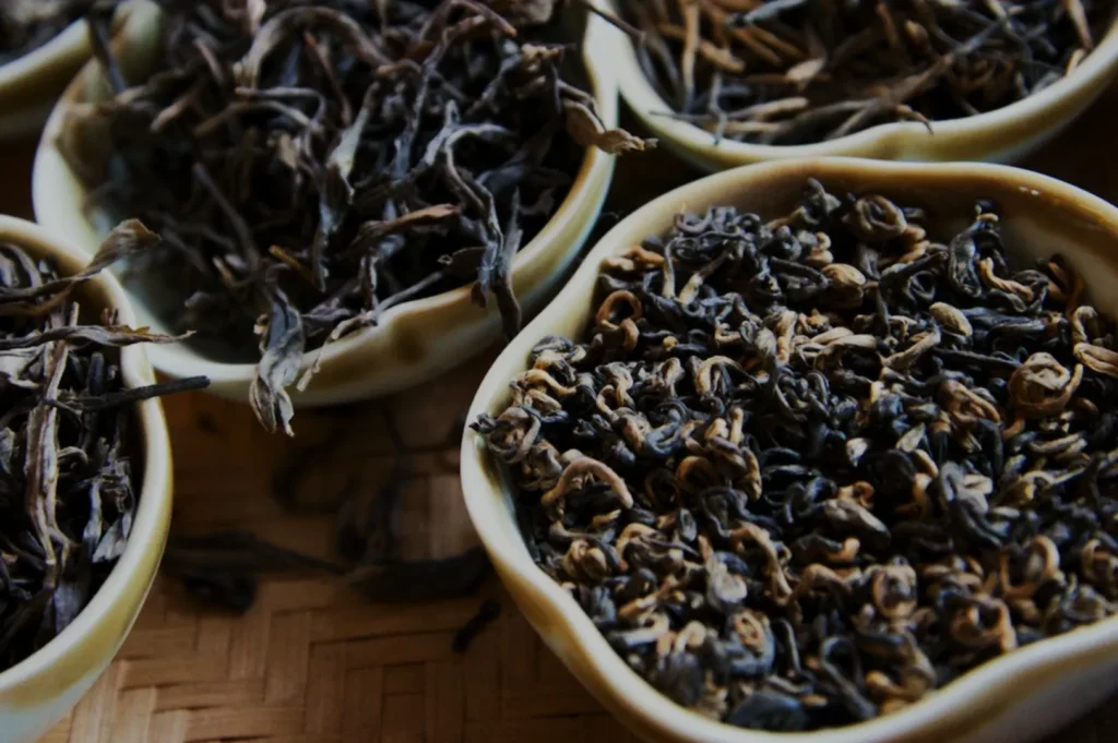 Close-up of dried tea leaves in ceramic bowls, showcasing assorted textures and dark hues. The mood is earthy and calming, evoking a sense of tradition.