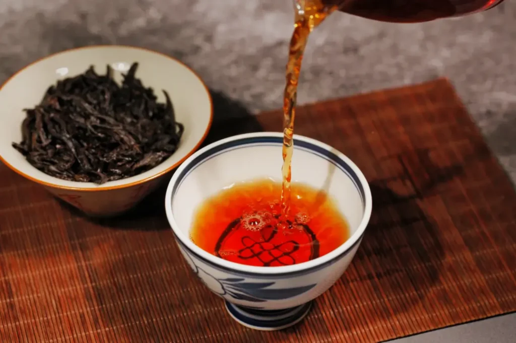Tea pouring into a patterned ceramic cup on a bamboo mat, beside a bowl of dry tea leaves; warm, inviting, and tranquil atmosphere.