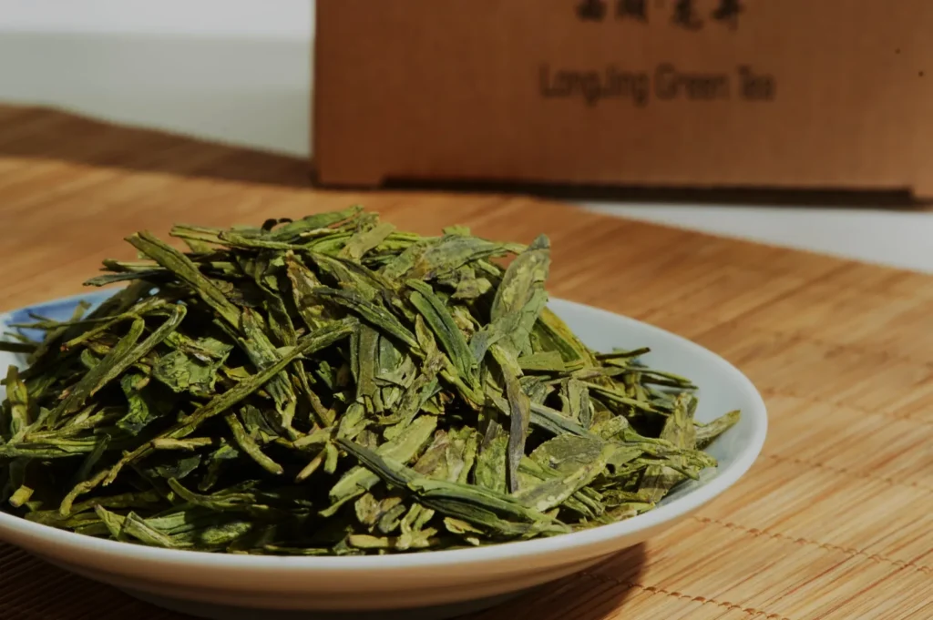 A plate of fresh green tea leaves on a bamboo mat, with a cardboard box labeled "Longjing Green Tea" in the background, conveying a sense of freshness.