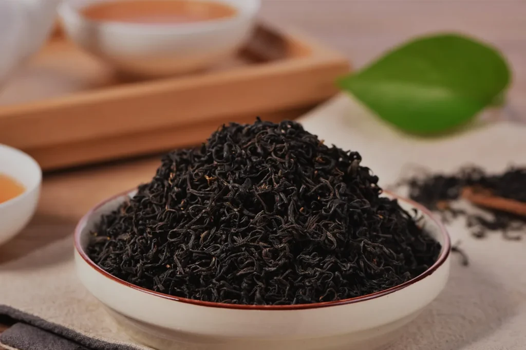 A bowl brimming with loose black tea leaves sits on a table, surrounded by cups of brewed tea. A green leaf adds a touch of freshness to the warm scene.