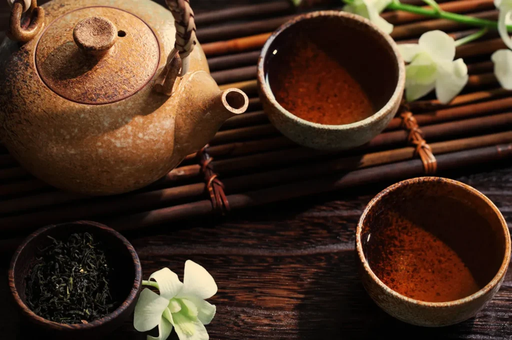A rustic teapot with two filled teacups on a bamboo mat, surrounded by loose tea leaves and white flowers. The scene conveys a calm, serene atmosphere.