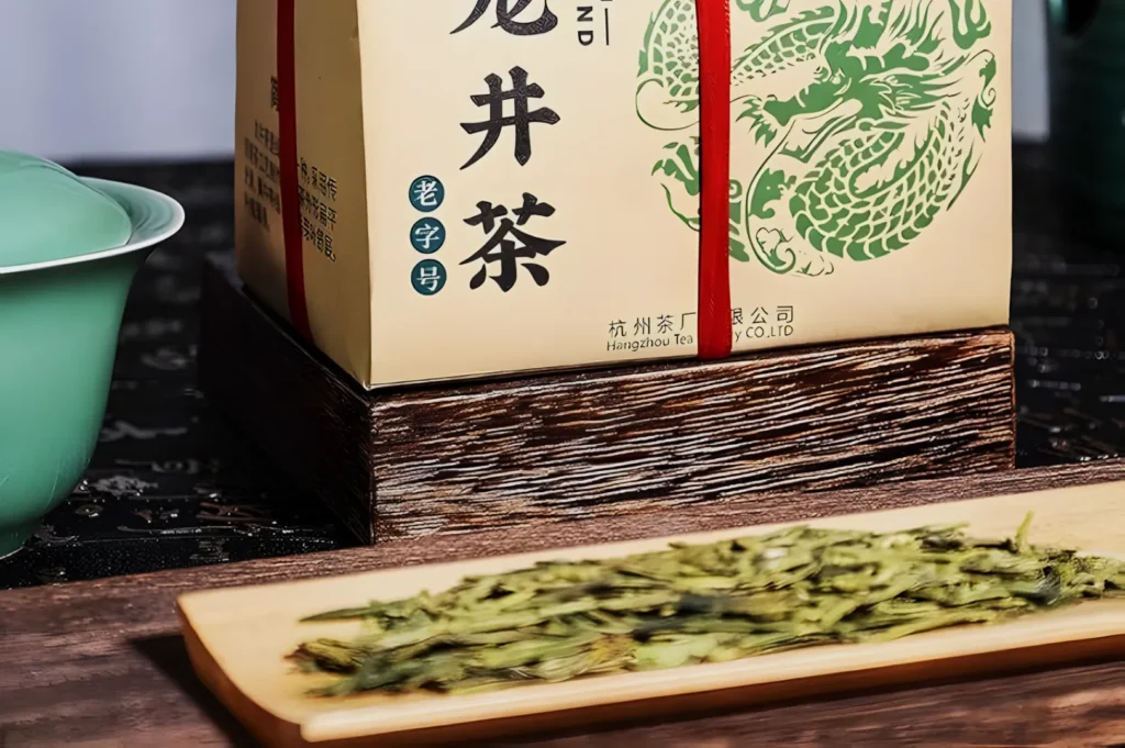 A box of Chinese tea with intricate green dragon design sits on a wooden base. In front, dried tea leaves are displayed on a wooden tray next to a green cup.