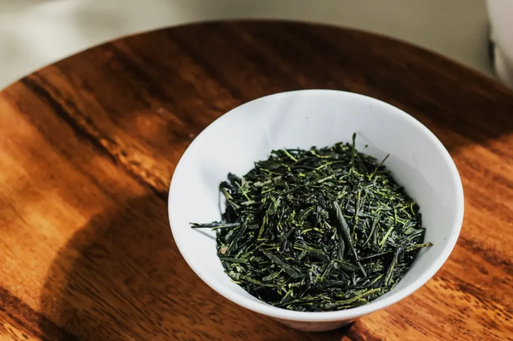 A white bowl filled with dried green tea leaves sits on a wooden table. Sunlight casts a soft shadow, creating a warm and serene atmosphere.