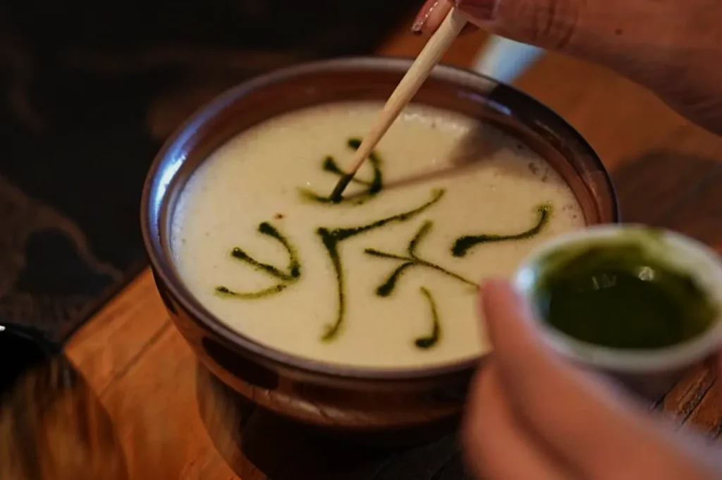 A person decorates creamy soup in a brown bowl with green sauce, creating abstract patterns using a thin stick, conveying a sense of artistry.
