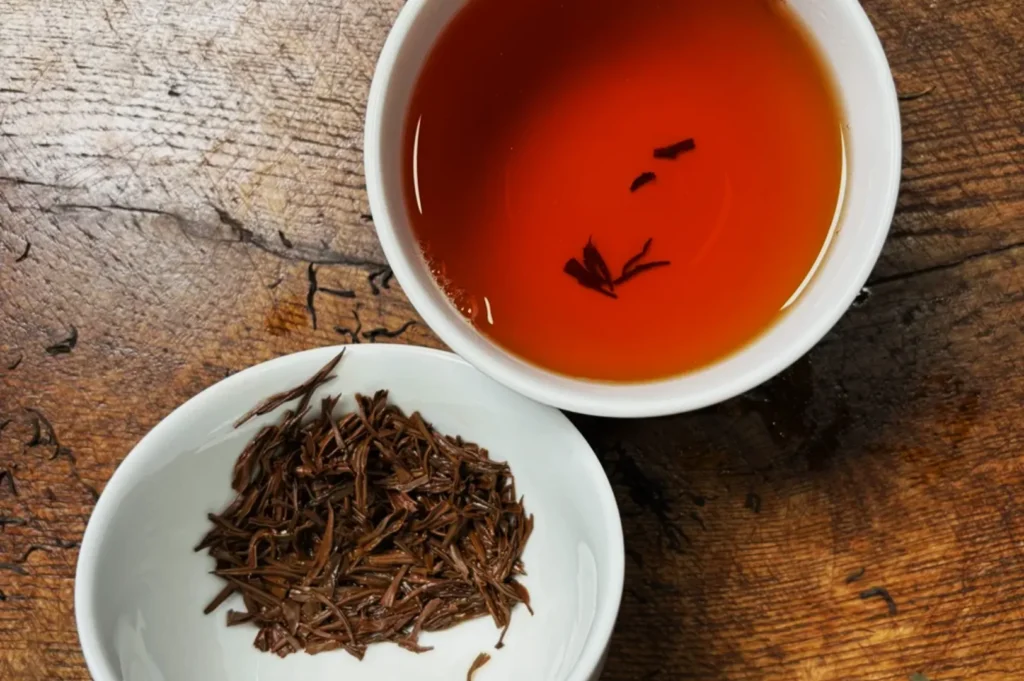 A white cup of amber tea with scattered leaves floats beside a small bowl of wet tea leaves on a rustic wooden table. Warm and inviting ambiance.