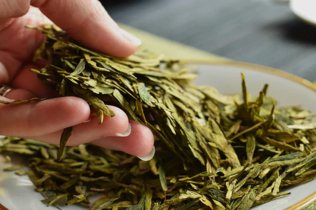 A hand gently holds a small pile of dried green tea leaves above a plate. The leaves are long and flat, creating a calm and natural tone.