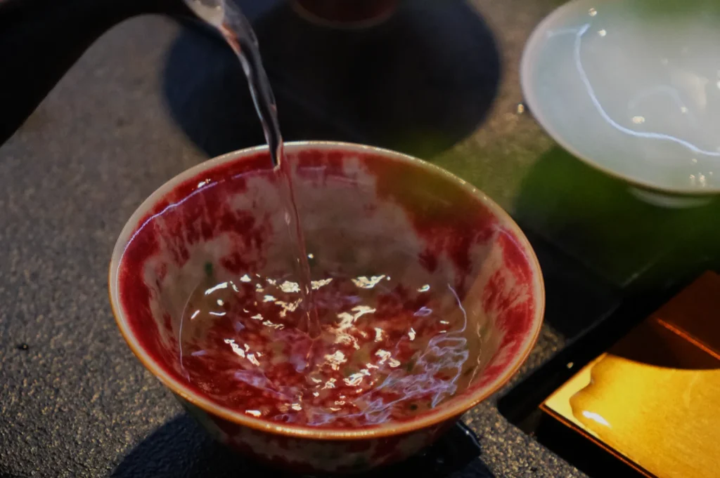 Pouring hot water into a red and white ceramic bowl, creating ripples. The setting conveys a warm, calming tea-making ritual.